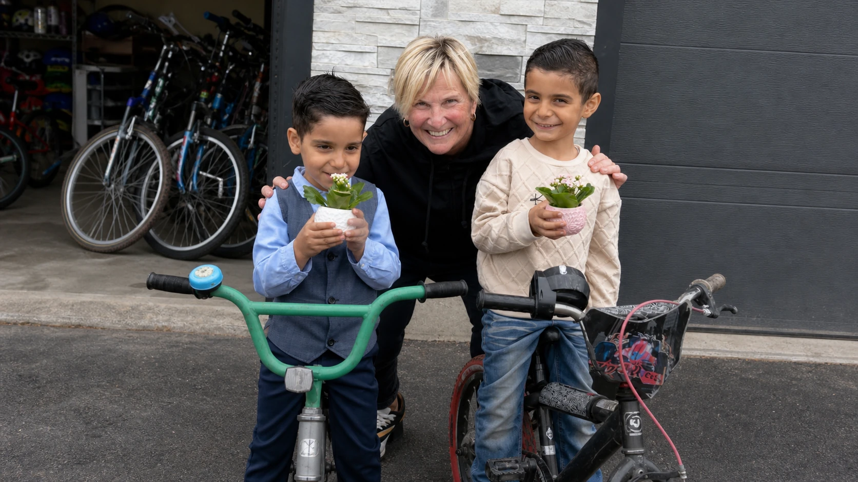 A candid community photo matching the source image: a Caucasian woman in her late 50s with light skin, short blonde hair, and a black jacket kneels between two Middle Eastern boys around 6 to 8 years old with olive skin, dark hair, and neat casual clothes. The boys stand beside children's bicycles and hold small potted plants, with a garage of repaired bikes in the background. No text overlay, no logo, natural documentary framing